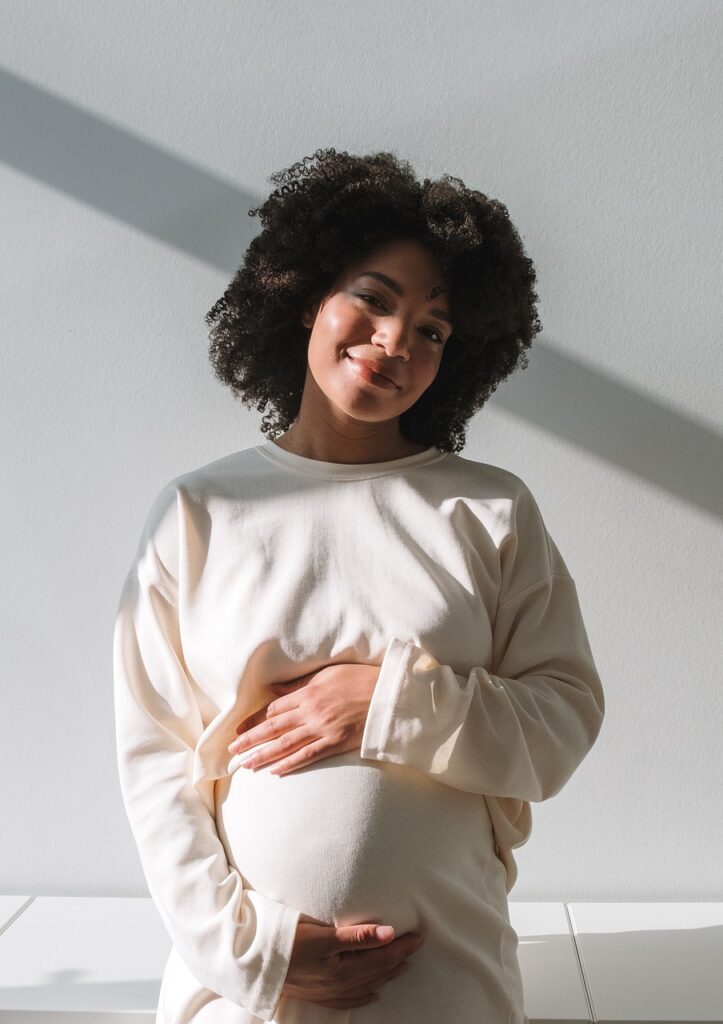 pregnant woman with left hand on top of pregnant belly and right hand under pregnant belly. head tipped to the left with a smile. Woman is wearing a white sweater .