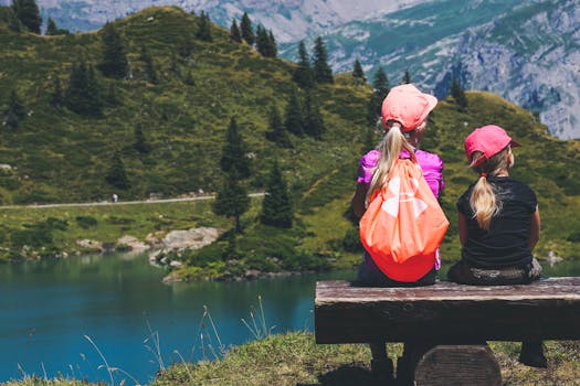 Two young girls sitting on a bench overlooking a serene mountain lake, enjoying a summer hike.